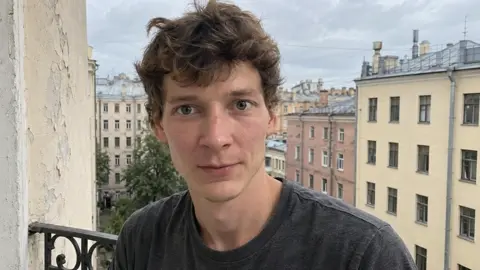 Парень на балконе чуть поворачивает голову к камере и мягко…
