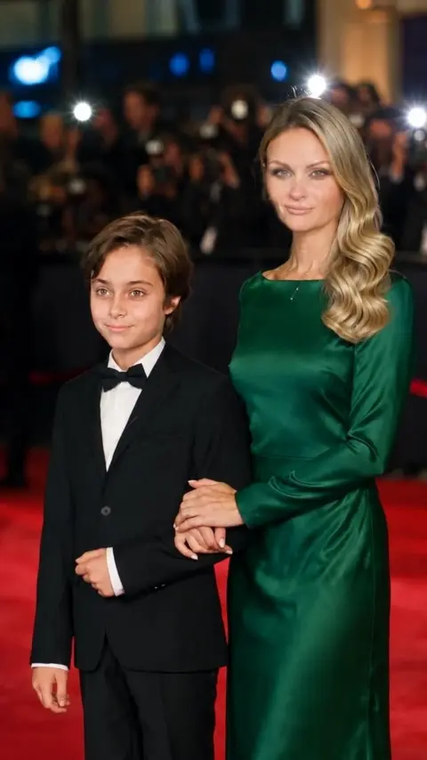 A red-carpet premiere at night. The boy in a tuxedo and his…